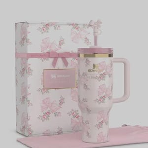 May include: A white Stanley Lovesha tumbler with a pink floral and bow pattern, a pink handle, and a matching lid and straw. The tumbler is presented with a matching gift box and a pink cloth.