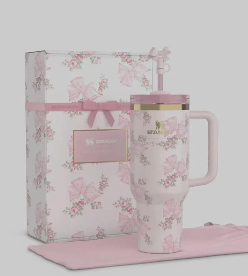 May include: A white Stanley Lovesha tumbler with a pink floral and bow pattern, a pink handle, and a matching lid and straw. The tumbler is presented with a matching gift box and a pink cloth.
