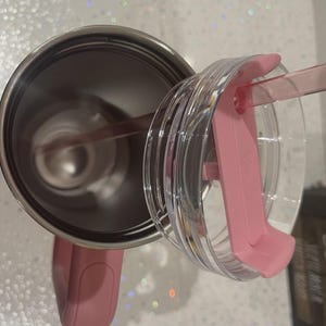 May include: A stainless steel tumbler with a pink handle and a matching pink lid. The lid is clear with a pink sliding closure and a pink straw inserted through the opening. The interior of the tumbler is metallic silver and hollow.