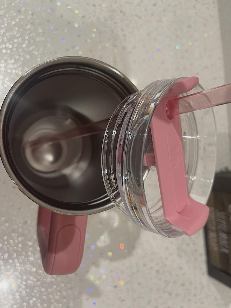 May include: A stainless steel tumbler with a pink handle and a matching pink lid. The lid is clear with a pink sliding closure and a pink straw inserted through the opening. The interior of the tumbler is metallic silver and hollow.