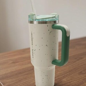May include: A cream-colored Stanley Starbucks tumbler with a speckled finish, a dark green handle, and a light green lid. The tumbler features a clear straw and the text Stanley + Starbucks printed vertically on the side of the cylindrical body.