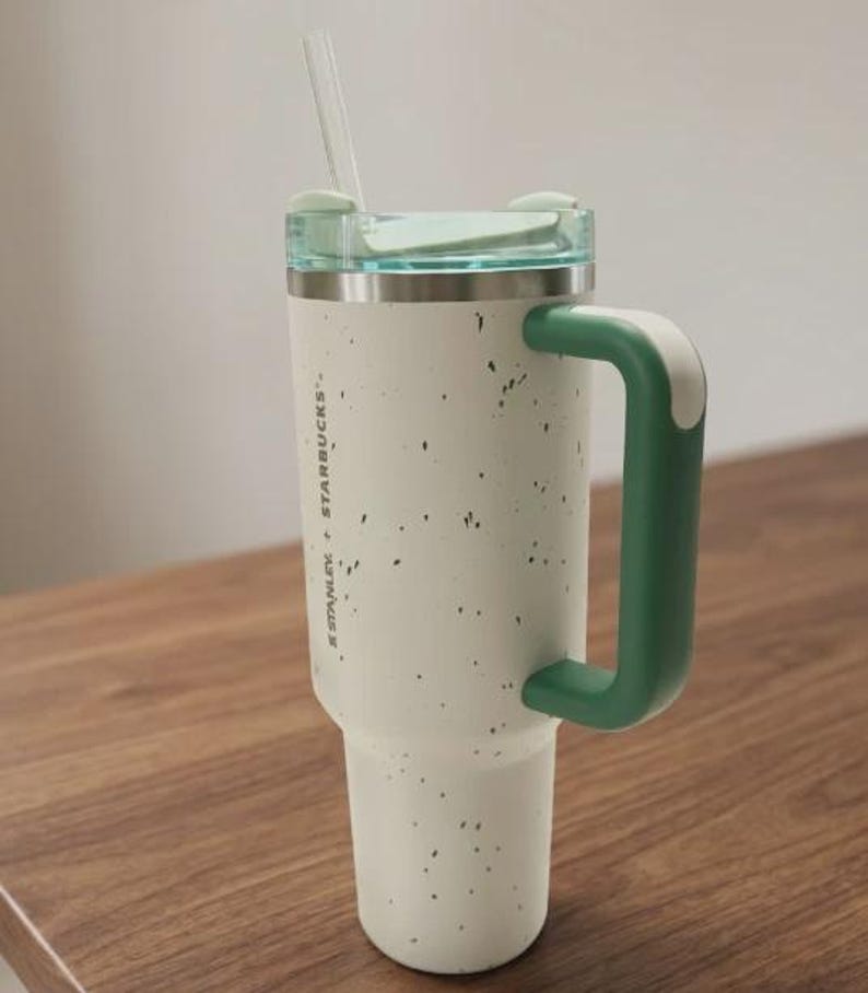 May include: A cream-colored Stanley Starbucks tumbler with a speckled finish, a dark green handle, and a light green lid. The tumbler features a clear straw and the text Stanley + Starbucks printed vertically on the side of the cylindrical body.