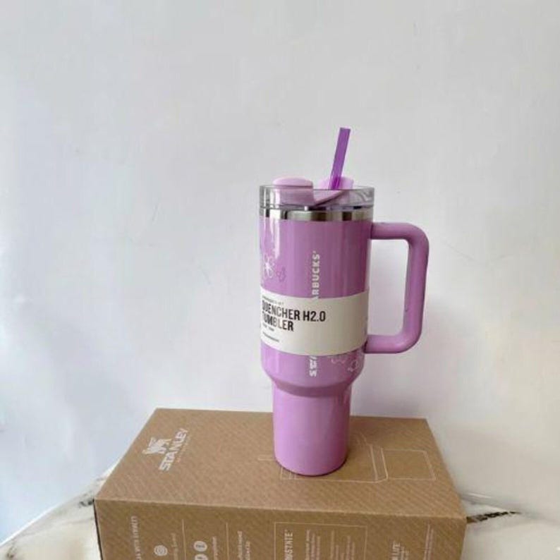 May include: A light purple Stanley Quencher H2.0 tumbler with a matching handle, a clear lid, and a purple straw. A white paper band wraps around the centre of the tumbler, featuring the text Starbucks and Quencher H2.0 Tumbler in black.