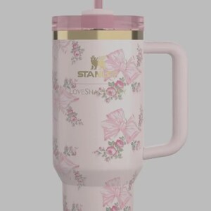 May include: A pink insulated travel mug with a handle, lid, and straw. The mug is adorned with pink bows and roses. A gold band accents the top. The text "STANLEY" and "LOVESHAPE" are printed on the mug.