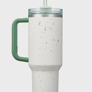 May include: A white stainless steel tumbler with a speckled pattern in dark green. The cup features a large, solid dark green handle on the side, a clear plastic lid, and a matching clear straw inserted through the center of the lid.