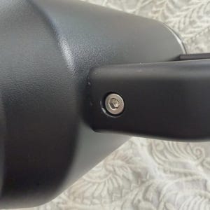 May include: Close-up of a black automotive side mirror. The mirror has a cylindrical shape and a rectangular arm. A silver bolt secures the arm to the mirror housing. The background is a patterned fabric.