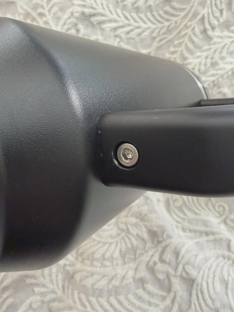 May include: Close-up of a black automotive side mirror. The mirror has a cylindrical shape and a rectangular arm. A silver bolt secures the arm to the mirror housing. The background is a patterned fabric.