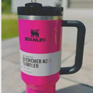 Op de afbeelding: Een felroze Stanley Quencher H2.0 Tumbler met een zwarte handgreep en deksel. Het heeft een doorzichtig rietje en het Stanley-logo. De tekst "QUENCHER H2.0 TUMBLER" staat op de zijkant.