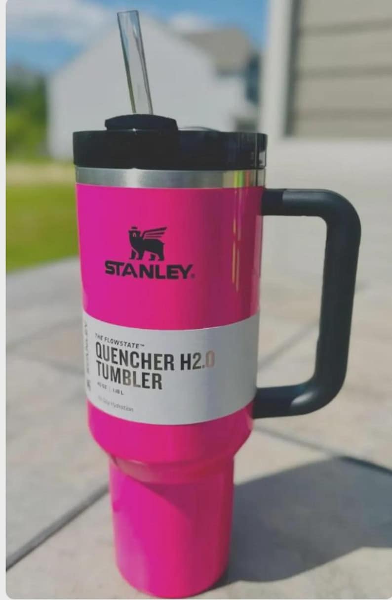 Op de afbeelding: Een felroze Stanley Quencher H2.0 Tumbler met een zwarte handgreep en deksel. Het heeft een doorzichtig rietje en het Stanley-logo. De tekst "QUENCHER H2.0 TUMBLER" staat op de zijkant.