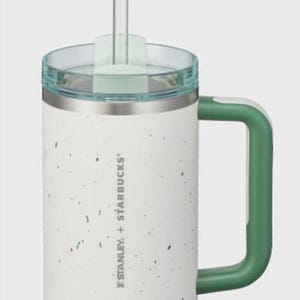 May include: A white Stanley + Starbucks tumbler with a speckled pattern, a green handle, and a clear lid with a matching green straw insert. The stainless steel rim sits below the lid, and the brand logo is printed vertically on the side.
