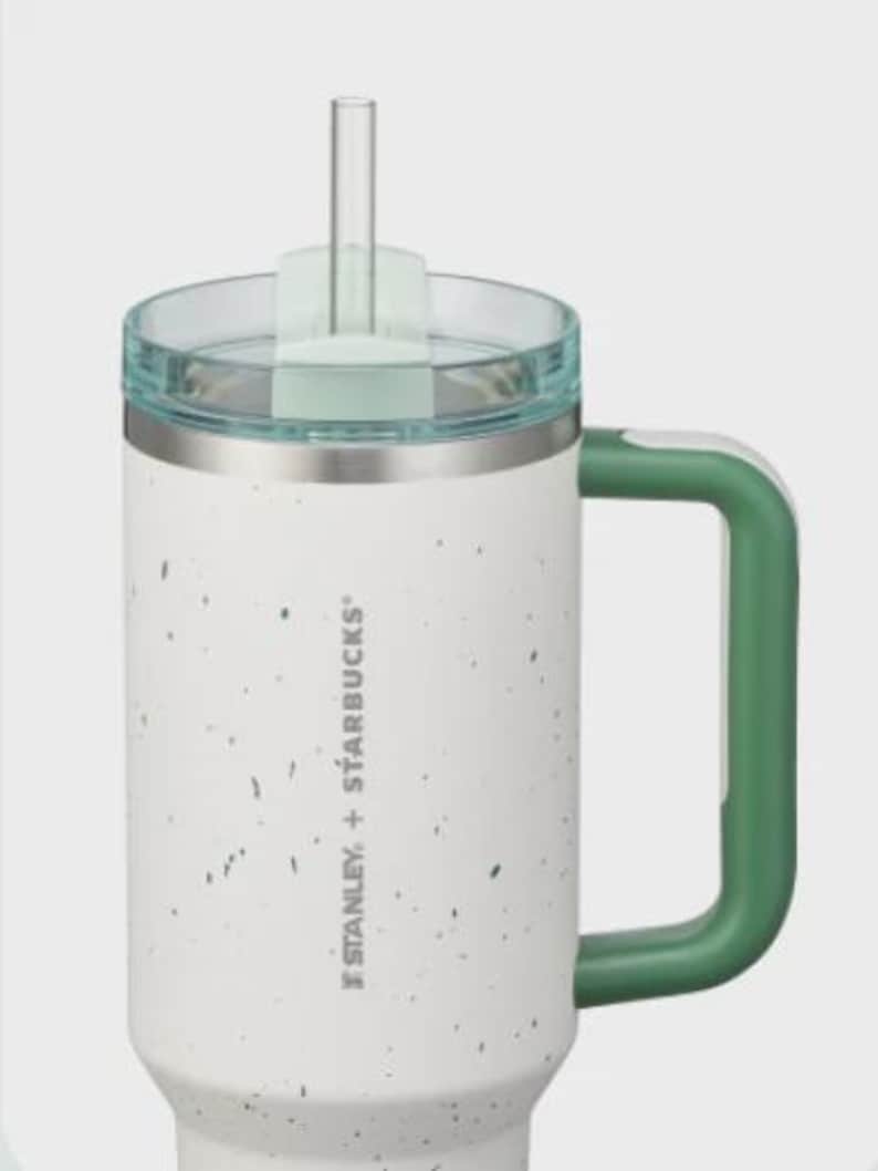 May include: A white Stanley + Starbucks tumbler with a speckled pattern, a green handle, and a clear lid with a matching green straw insert. The stainless steel rim sits below the lid, and the brand logo is printed vertically on the side.