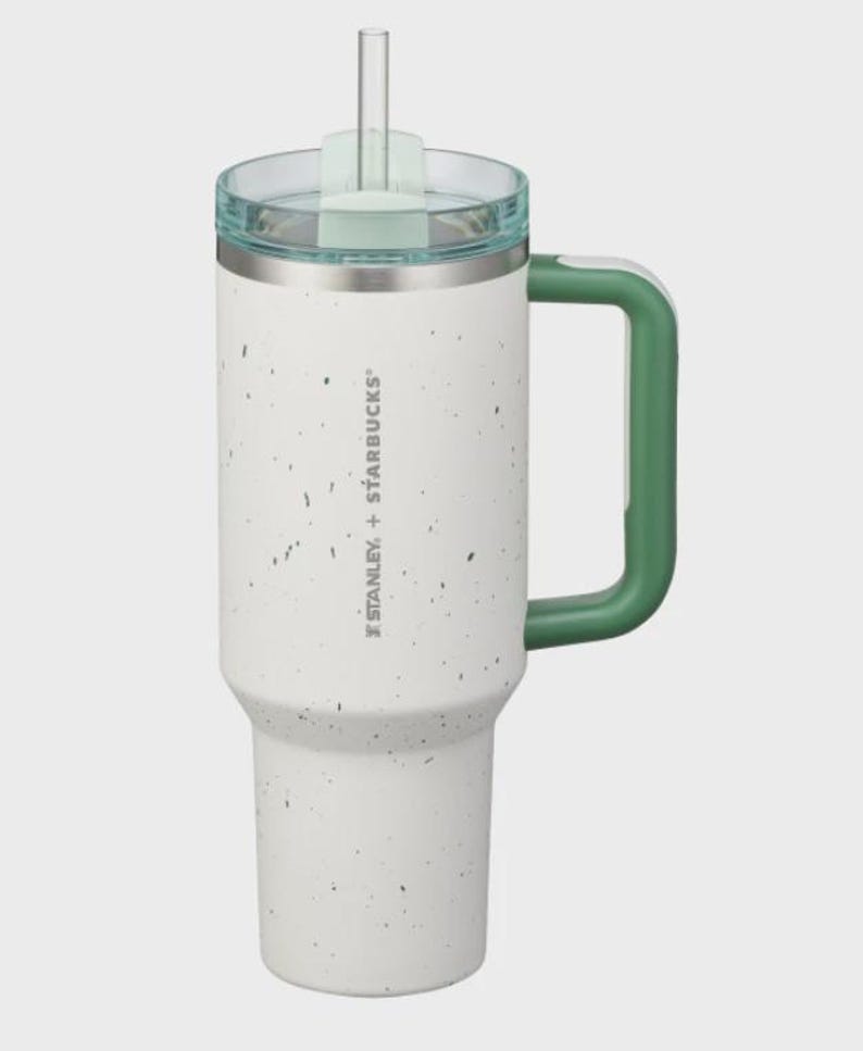 May include: A white tumbler with a speckled finish, a green handle, and a clear lid with a straw. The side features the text STANLEY + STARBUCKS. The cup has a tapered base and a cylindrical body, designed for holding beverages with a secure lid.