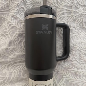 May include: Black Stanley Quencher H2.0 tumbler with a handle and straw. The tumbler has the Stanley logo and the text "QUENCHER TUMBLER H2.0" on the side. It holds 1.18L.