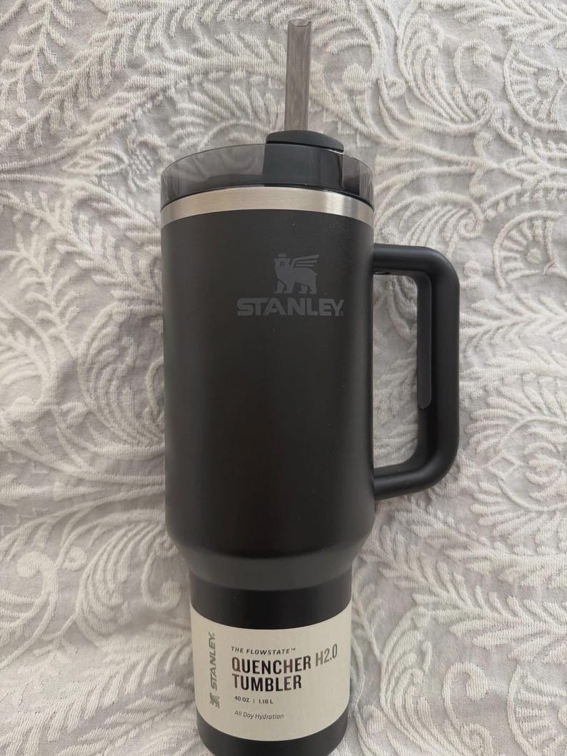 May include: Black Stanley Quencher H2.0 tumbler with a handle and straw. The tumbler has the Stanley logo and the text "QUENCHER TUMBLER H2.0" on the side. It holds 1.18L.