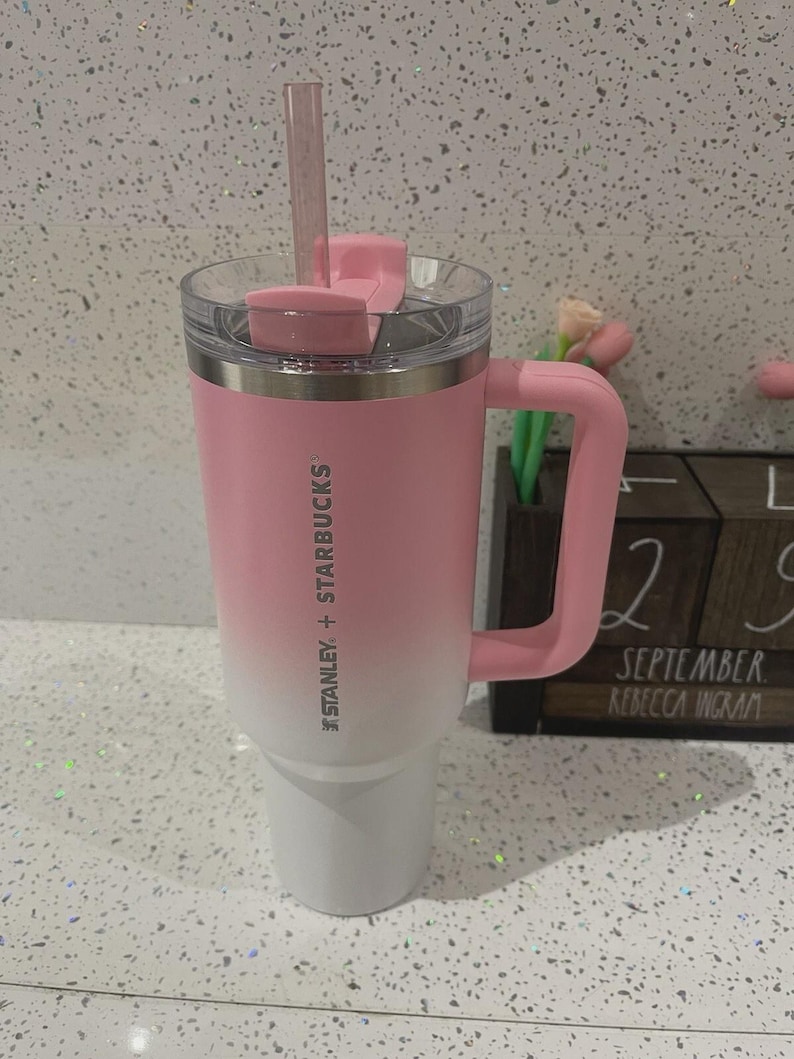 May include: A Stanley + Starbucks tumbler featuring a pink to white ombre finish, a matching pink handle, a clear lid with a pink rotating cover, and a clear straw. The branding is printed vertically in white on the side of the stainless steel cup.