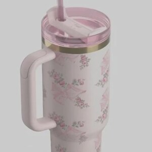 May include: A light pink insulated tumbler with a handle, straw, and lid. The tumbler features a floral and umbrella pattern in pink and white. A gold band accents the top.