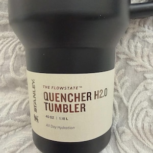 May include: Black Stanley Quencher H2.0 tumbler with a handle. The tumbler has a white label with the text "THE FLOWSTATE", "QUENCHER H2.0 TUMBLER", "40 OZ | 1.18 L", and "All Day Hydration".