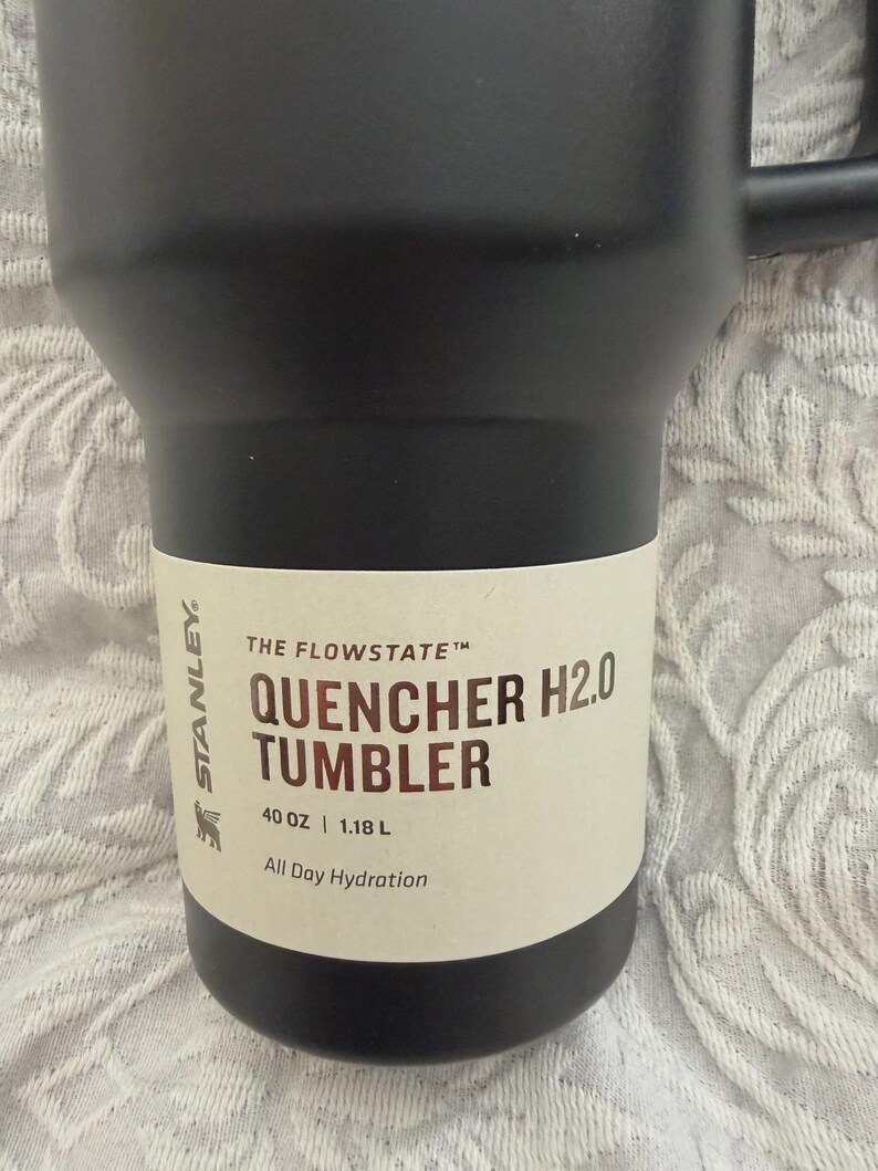 May include: Black Stanley Quencher H2.0 tumbler with a handle. The tumbler has a white label with the text "THE FLOWSTATE", "QUENCHER H2.0 TUMBLER", "40 OZ | 1.18 L", and "All Day Hydration".