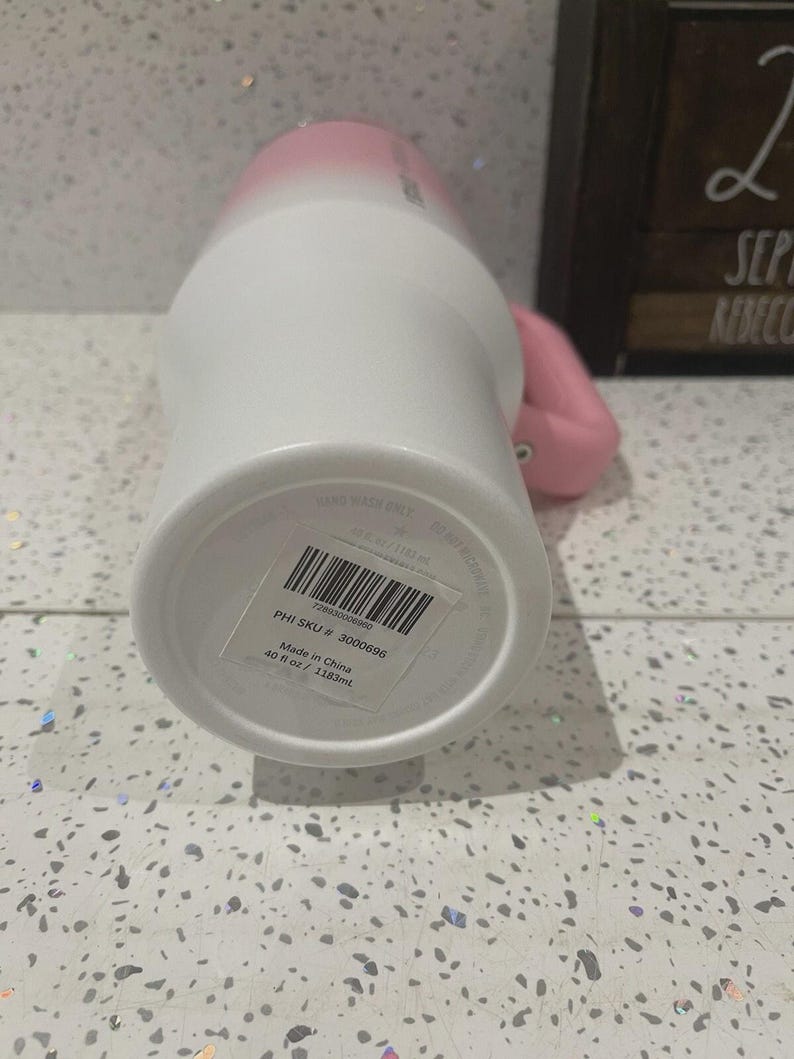 May include: A white and pink ombre tumbler with a handle, featuring a sticker on the base with a barcode, the text PHI SKU # 3000696, and the capacity of 40 fl oz / 1183 ml. The tumbler has a cylindrical body and a sturdy side handle.