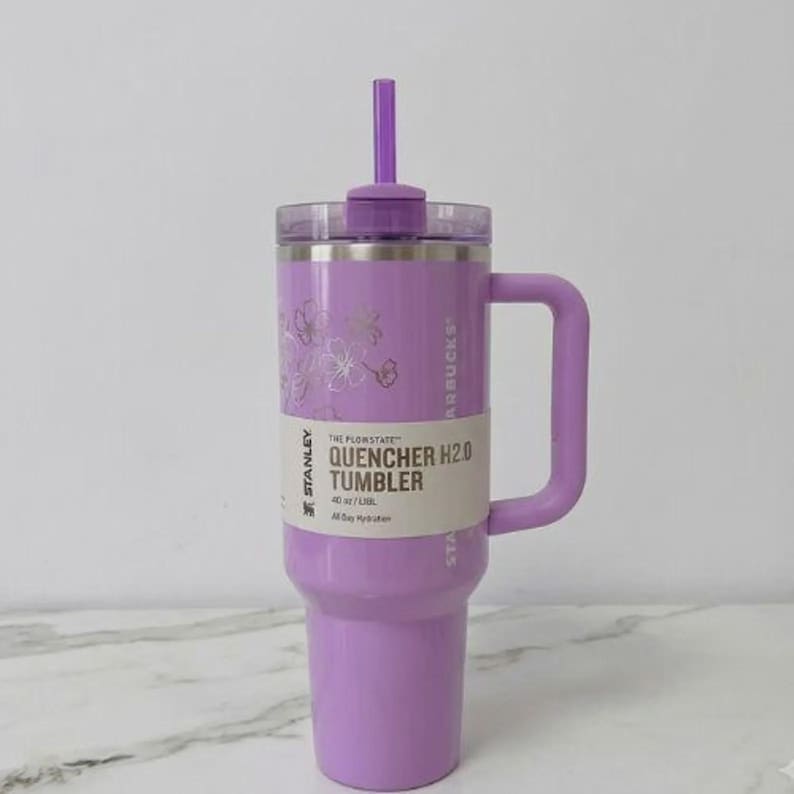 May include: A light purple Stanley Quencher H2.0 tumbler with a 1.18 litre capacity, featuring a matching purple straw, a side handle, and a clear lid. A paper band around the centre displays the text Stanley, The Flowstate, and Quencher H2.0 Tumbler.