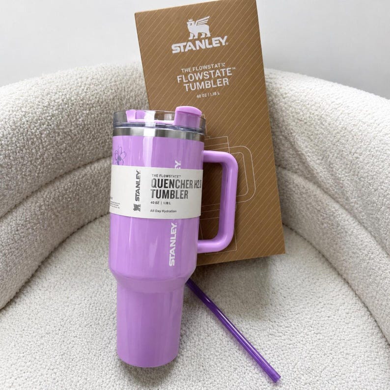 May include: A light purple Stanley Flowstate Quencher H2.0 tumbler with a 1.18 L capacity, a matching handle, a clear lid, and a purple straw. The tumbler features a white product label and is positioned in front of its original brown cardboard box.