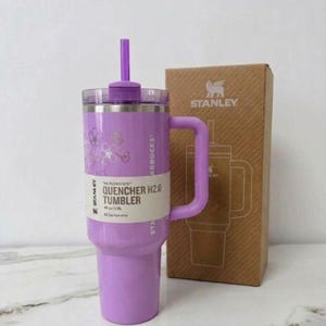 May include: A light purple Stanley Quencher H2.0 tumbler with a matching straw and handle. The tumbler features a white label with the text "THE FLOWSTATE QUENCHER H2.0 TUMBLER 40 OZ/1.18L". A brown cardboard box with the Stanley logo is positioned behind it.