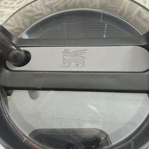 May include: A close-up shot of a clear, circular lid with a black rim and handle. A grey rectangular bar with a winged bear logo is centered on the lid. The lid appears to be made of durable plastic.