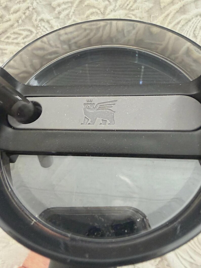 May include: A close-up shot of a clear, circular lid with a black rim and handle. A grey rectangular bar with a winged bear logo is centered on the lid. The lid appears to be made of durable plastic.
