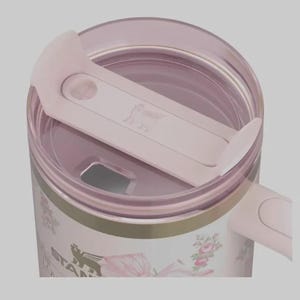 May include: A close-up of a pink and white floral Stanley tumbler with a matching pink lid. The lid has a sliding closure and the Stanley logo is visible. The tumbler has a gold band near the top and floral designs.