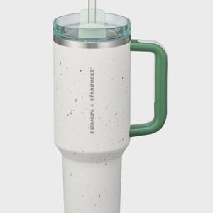 May include: A white tumbler with a speckled finish, a green handle, and a clear lid with a straw. The side features the text STANLEY + STARBUCKS. The cup has a tapered base and a cylindrical body, designed for holding beverages with a secure lid.