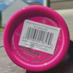 Op de afbeelding: Close-up van de bodem van een felroze Stanley-beker. Een witte sticker met een barcode en productinformatie is bevestigd. De tekst "STANLEY" is zichtbaar.