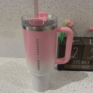 May include: A Stanley + Starbucks tumbler featuring a pink to white ombre finish, a matching pink handle, a clear lid with a pink rotating cover, and a clear straw. The branding is printed vertically in white on the side of the stainless steel cup.