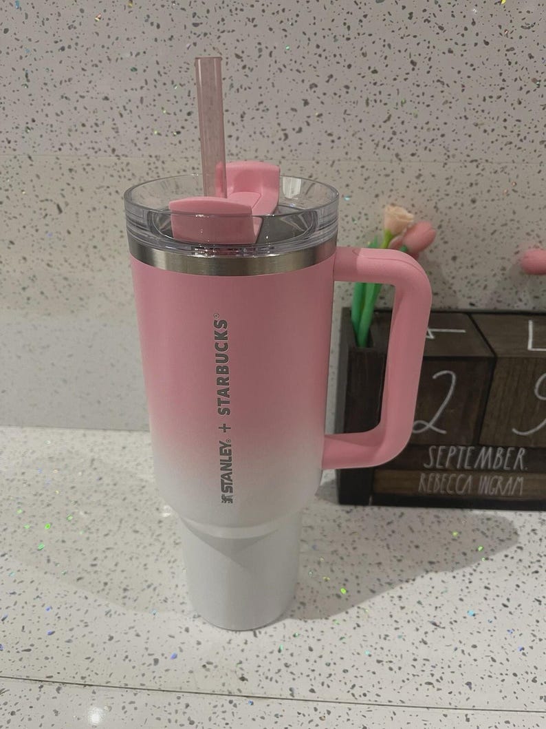 May include: A Stanley + Starbucks tumbler featuring a pink to white ombre finish, a matching pink handle, a clear lid with a pink rotating cover, and a clear straw. The branding is printed vertically in white on the side of the stainless steel cup.