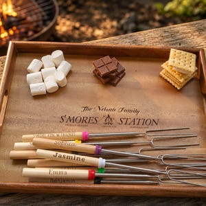 May include: A rectangular wooden serving tray engraved with The Nelson Family S'MORES STATION, containing white marshmallows, chocolate squares, graham crackers, and several metal roasting sticks with personalized wooden handles.