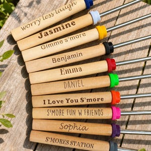 Puede incluir: Un juego de diez brochetas de metal con mangos de madera clara. Cada mango está grabado con texto que incluye "worry less laugh", "Jasmine", "Gimme s'more", "Benjamin", "Emma", "DANIEL", "I Love You S'more", "S'MORE FUN W FRIENDS", "Sophia" y "S'MORES STATION". Cada brocheta tiene un anillo de plástico de color único en la base del mango.