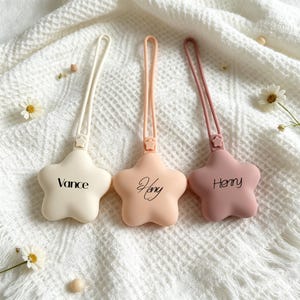 May include: Three star-shaped silicone pacifier clips with loops, each featuring a small star accent at the base of the strap. The clips are colored cream, peach, and dusty rose, with the names Vance, Heny, and Henry printed in black script.