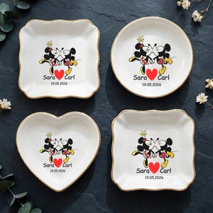 Op de afbeelding: Vier witte keramische schalen met gouden randen, elk met een Mickey en Minnie Mouse ontwerp met een rood hart en de namen "Sara" en "Carl" met de datum "19.05.2026". De schalen zijn vierkant, rond, hartvormig en vierkant met geschulpte rand.