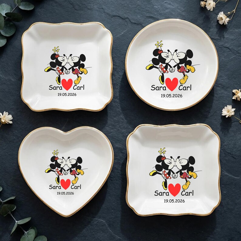 Op de afbeelding: Vier witte keramische schalen met gouden randen, elk met een Mickey en Minnie Mouse ontwerp met een rood hart en de namen "Sara" en "Carl" met de datum "19.05.2026". De schalen zijn vierkant, rond, hartvormig en vierkant met geschulpte rand.