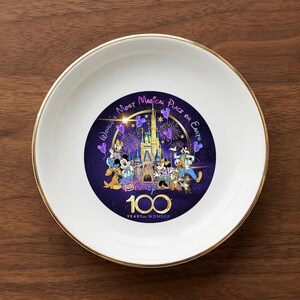 Op de afbeelding: Een witte keramische schaal met een gouden rand met een Disney 100 Years of Wonder-ontwerp. Het ontwerp bevat Disney-personages, een kasteel en de tekst "World's Most Magical Place on Earth."