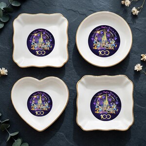 Op de afbeelding: Vier witte keramische schaaltjes met gouden randen. Elk schaaltje heeft een centraal ontwerp met Disney-personages, een kasteel en de tekst "100 Years of Wonder". De schaaltjes zijn in vierkante, ronde, hart- en geschulpte vierkante vormen.