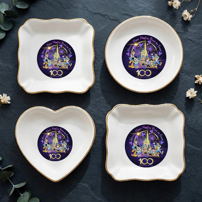 Op de afbeelding: Vier witte keramische schaaltjes met gouden randen. Elk schaaltje heeft een centraal ontwerp met Disney-personages, een kasteel en de tekst "100 Years of Wonder". De schaaltjes zijn in vierkante, ronde, hart- en geschulpte vierkante vormen.