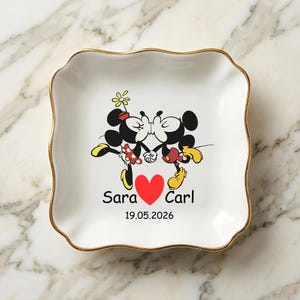 Op de afbeelding: Witte keramische sieradenschaal met een gouden rand en een golvende rand. De schaal heeft een cartoonafbeelding van Mickey en Minnie Mouse die kussen, met een rood hart en de namen "Sara" en "Carl" en de datum "19.05.2026".