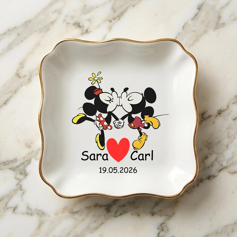 Op de afbeelding: Witte keramische sieradenschaal met een gouden rand en een golvende rand. De schaal heeft een cartoonafbeelding van Mickey en Minnie Mouse die kussen, met een rood hart en de namen "Sara" en "Carl" en de datum "19.05.2026".