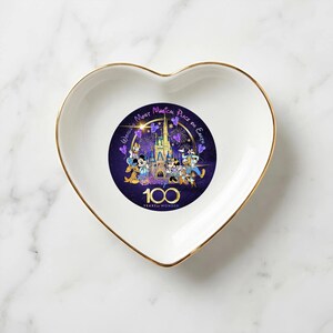 Op de afbeelding: Wit hartvormig sieradenschaaltje met een gouden rand. Het midden heeft een Disney-ontwerp met personages, een kasteel en de tekst "World's Most Magical Place on Earth" en "100 Years of Wonder".