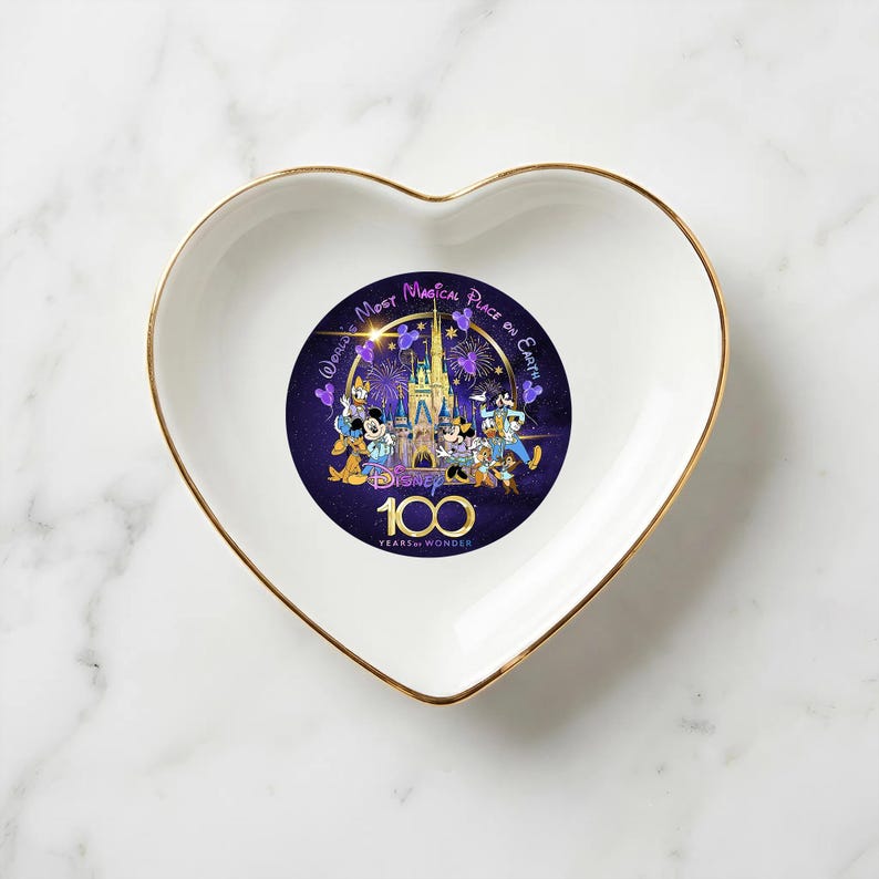 Op de afbeelding: Wit hartvormig sieradenschaaltje met een gouden rand. Het midden heeft een Disney-ontwerp met personages, een kasteel en de tekst "World's Most Magical Place on Earth" en "100 Years of Wonder".