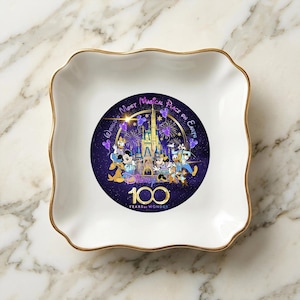 Op de afbeelding: Een witte keramische sieradenschaal met een golvende rand en gouden rand. Het midden heeft een Disney 100 Years of Wonder-ontwerp met personages en een kasteel op een donkerpaarse achtergrond. De schaal is vierkant met afgeronde hoeken.