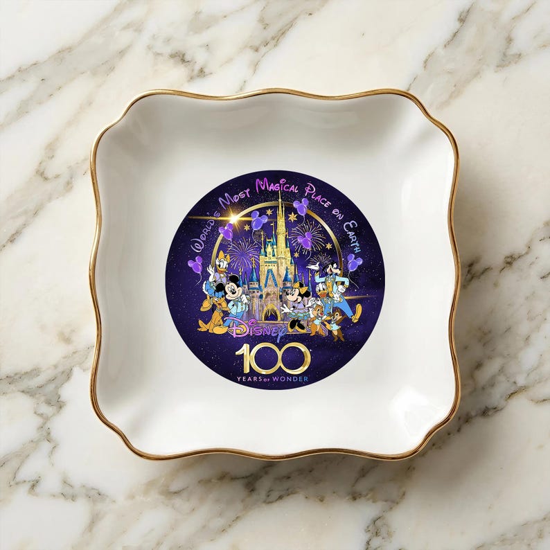 Op de afbeelding: Een witte keramische sieradenschaal met een golvende rand en gouden rand. Het midden heeft een Disney 100 Years of Wonder-ontwerp met personages en een kasteel op een donkerpaarse achtergrond. De schaal is vierkant met afgeronde hoeken.