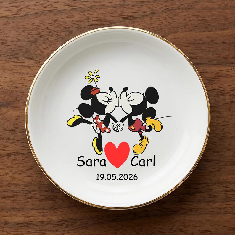 Op de afbeelding: Witte keramische plaat met een gouden rand met een cartoonafbeelding van Mickey en Minnie Mouse die kussen. Een rood hart is gecentreerd onder de karakters, met de namen "Sara" en "Carl" aan weerszijden. De datum "19.05.2026" staat eronder gedrukt.