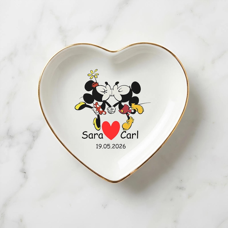 Op de afbeelding: Wit hartvormig schaaltje met een gouden rand. Het heeft een cartoonafbeelding van Mickey en Minnie Mouse die kussen, met een rood hart en de namen "Sara" en "Carl" en de datum "19.05.2026".