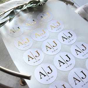 Könnte beinhalten: Ein Bogen mit runden weißen Aufklebern, die ein Goldfolien-Monogramm mit den Buchstaben A und J zeigen, getrennt durch eine vertikale Linie. Unter dem Monogramm ist auf jedem Aufkleber das Datum 02.08.2027 in kleiner, schwarzer Schrift gedruckt.